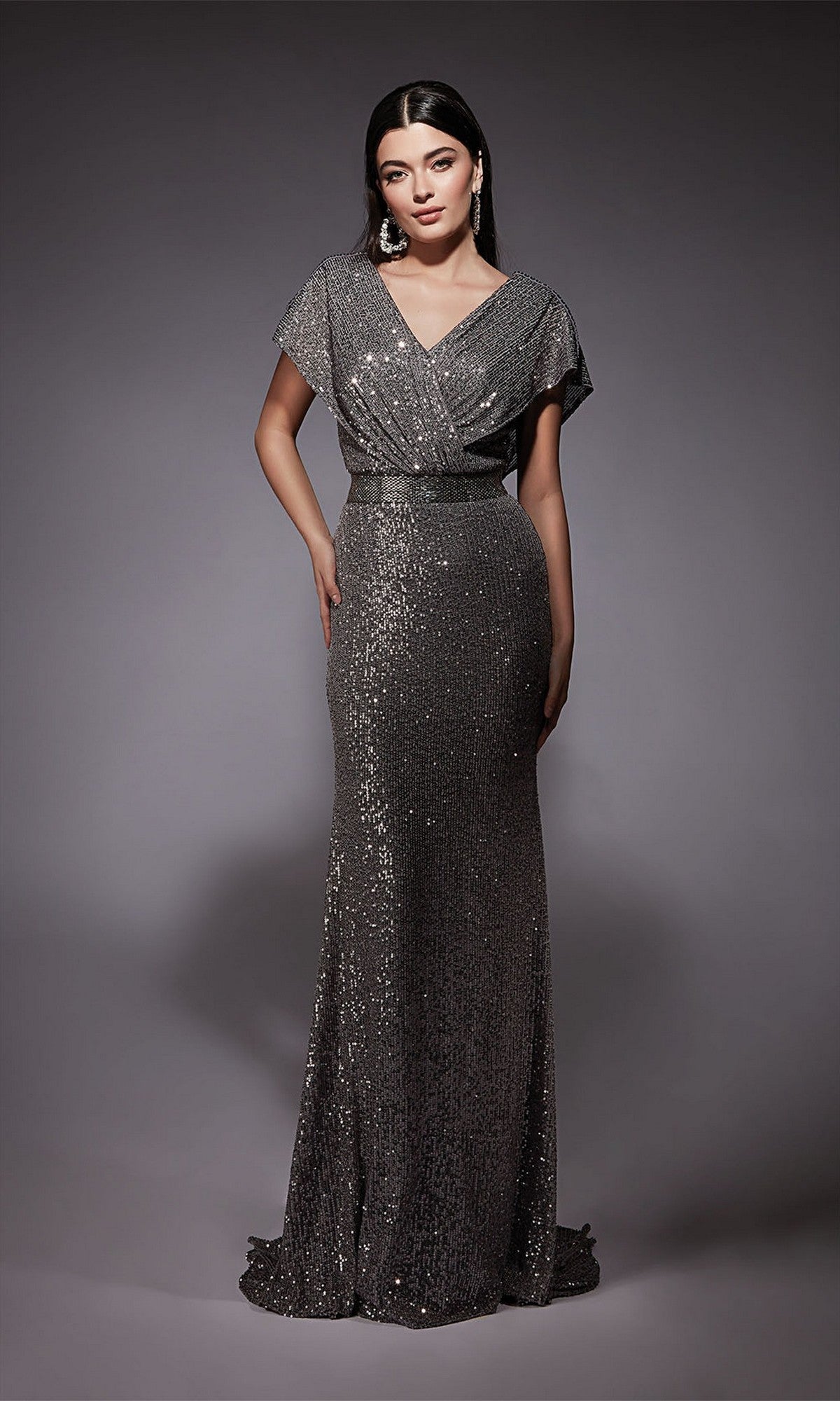 A woman with long dark hair and earrings stands against a gray backdrop, wearing the gunmetal Alyce 27708 long formal dress with short sleeves, a v-neckline, fitted waist, beaded waistband, and shimmering sequins.